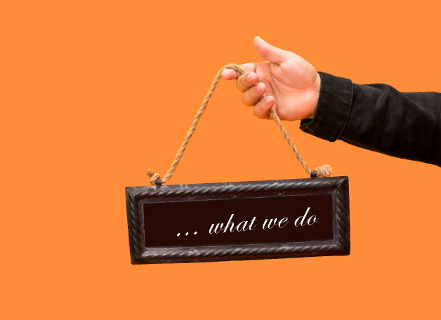 Hand holding a small black sign with white text reading '... what we do' against an orange background, representing services provided by TechnicaX, a Microsoft Dynamics 365 solutions provider in the USA. 